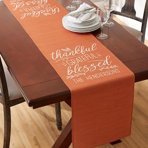 May include: An orange table runner with the words "thankful, grateful, blessed" in white script. The runner is placed on a dark brown wooden table, with white plates, silverware, and wine glasses.