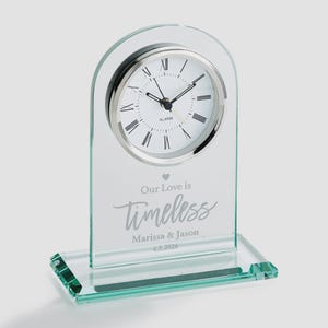 Op de afbeelding: Een heldere glazen wekker met een zilverkleurige wijzerplaat en Romeinse cijfers. De klok heeft de woorden "Our Love is Timeless" en de namen "Marissa & Jason" met de datum "4.8.2026" gegraveerd.