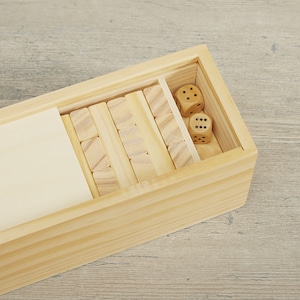 Puede incluir: Un juego de madera con una caja rectangular que contiene bloques de madera y dos dados. Los bloques est&aacute;n dispuestos en una estructura similar a una torre, y los dados muestran diferentes n&uacute;meros de puntos. La caja es de madera clara.
