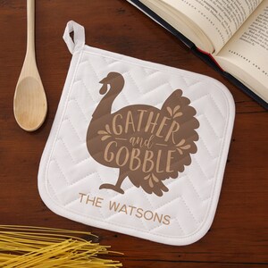 May include: White quilted pot holder with a brown silhouette of a turkey with the text "Gather and Gobble" and the name "The Watsons" printed below.