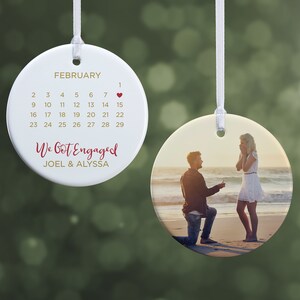 May include: A white ceramic ornament with a calendar showing February and the date of the engagement. The ornament features the text "We Got Engaged" and the names "JOEL & ALYSSA". The other side shows a photo of a proposal.