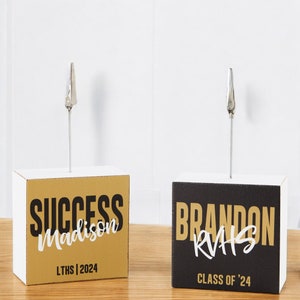 May include: Two white photo holders with gold and black accents. The holder on the left has a gold background with black text that says "SUCCESS Madison LTHS | 2024". The holder on the right has a black background with gold text that says "BRANDON RVAS CLASS OF '24".