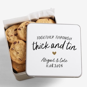 May include: A metal tin filled with chocolate chip cookies. The tin has a white lid with the text "TOGETHER THROUGH thick and tin" and the names Abigail & Cole with the date 11.08.2025.