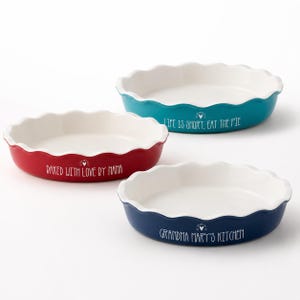 May include: Three ceramic pie dishes in red, teal, and navy blue. Each dish has a white interior and a scalloped edge. Text on the dishes reads "BAKED WITH LOVE BY NANA", "LIFE IS SHORT, EAT THE PIE", and "GRANDMA MARY'S KITCHEN".