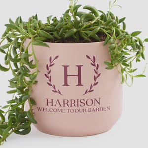 May include: A light pink ceramic planter with a trailing green succulent. The planter features the name "HARRISON" and the phrase "WELCOME TO OUR GARDEN" in a dark purple font, along with the letter "H" inside a wreath.