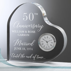 May include: A heart-shaped, clear glass anniversary clock. The clock face is round with Roman numerals. The text on the glass reads "50th Anniversary," names, the date "June 15, 1974," and "Until the end of time..."