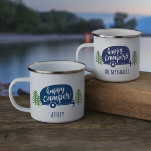 May include: Two white enamel mugs with silver rims featuring a blue camper graphic, green pine trees, and the text "happy camper" with the name "ASHLEY" on one, and "happy campers" with "THE MARSHALLS" on the other, set on a wooden surface.