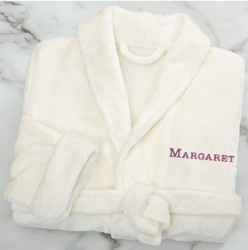 Peut inclure: Peignoir blanc en peluche avec col ch&acirc;le et ceinture &agrave; nouer. Le peignoir est brod&eacute; du nom "MARGARET" sur le devant en fil violet.