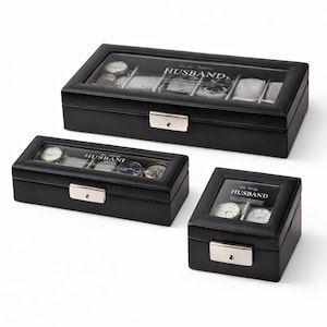 May include: Three black watch display cases with glass tops. The cases have silver clasps and the text "For My Dapper Husband" is visible. The cases are rectangular and square, and are designed to hold watches.