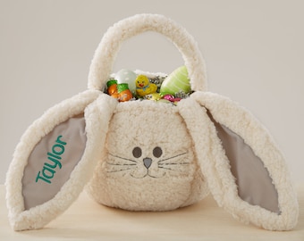 May include: A plush, cream-colored Easter basket shaped like a bunny. The basket has long, floppy ears with gray lining and the name "Taylor" embroidered in green. The basket is filled with Easter eggs and treats.