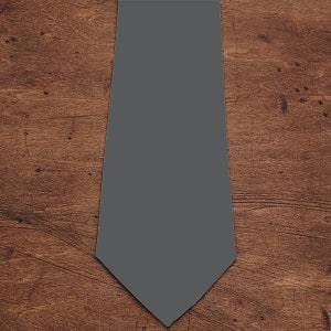 May include: A plain grey tie, presented on a wooden surface. The tie is a mid-tone grey, with a traditional pointed tip. The background features a warm-toned, textured wood grain.