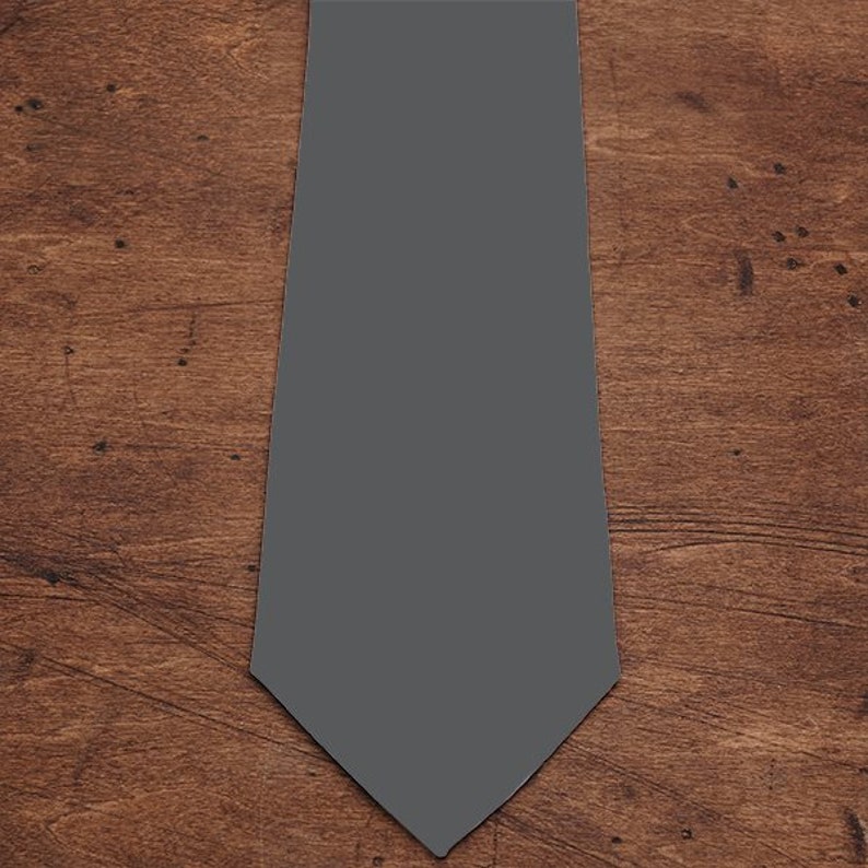 May include: A plain grey tie, presented on a wooden surface. The tie is a mid-tone grey, with a traditional pointed tip. The background features a warm-toned, textured wood grain.
