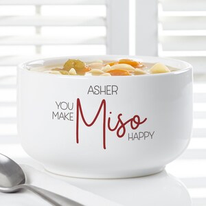 K&ouml;nnte beinhalten: Eine wei&szlig;e Keramik-Suppensch&uuml;ssel gef&uuml;llt mit Suppe, mit Karotten, Nudeln und Gem&uuml;se. Die Sch&uuml;ssel tr&auml;gt den Text "YOU MAKE Miso HAPPY" mit dem Namen "ASHER" dar&uuml;ber. Ein silberner L&ouml;ffel ist sichtbar.