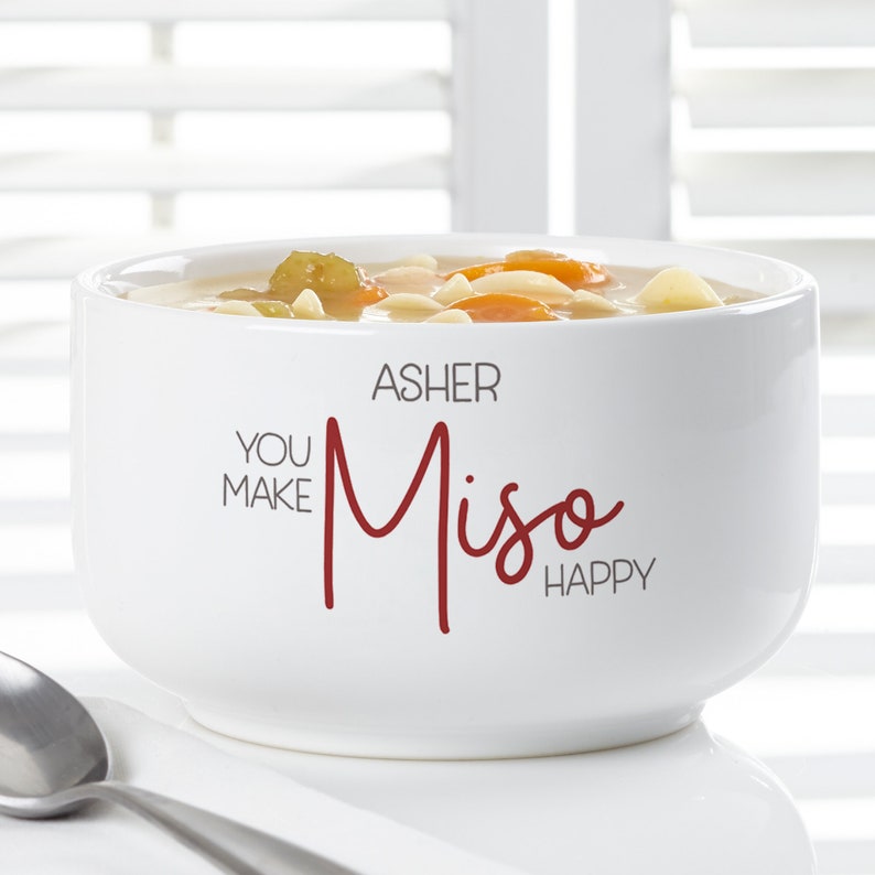K&ouml;nnte beinhalten: Eine wei&szlig;e Keramik-Suppensch&uuml;ssel gef&uuml;llt mit Suppe, mit Karotten, Nudeln und Gem&uuml;se. Die Sch&uuml;ssel tr&auml;gt den Text "YOU MAKE Miso HAPPY" mit dem Namen "ASHER" dar&uuml;ber. Ein silberner L&ouml;ffel ist sichtbar.