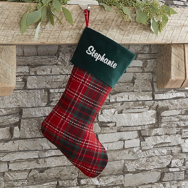 May include: A Christmas stocking with a red and green plaid pattern. The cuff is dark green with the name "Stephanie" embroidered in white. The stocking hangs from a wooden beam.