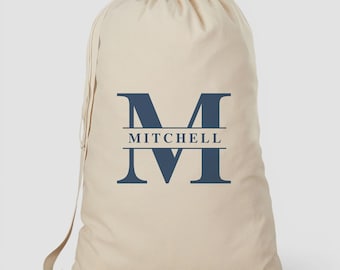 May include: A beige drawstring laundry bag featuring a large navy blue letter M with the name MITCHELL printed in navy blue capital letters across the centre of the letter. The bag has a cinched top closure with a grey fabric drawstring cord.