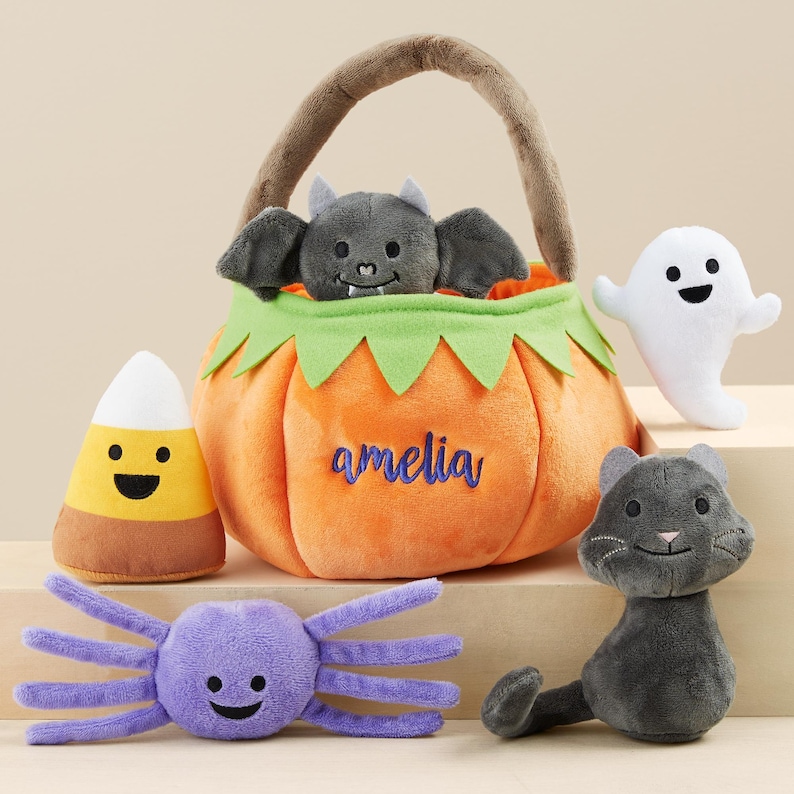 K&ouml;nnte beinhalten: Eine Sammlung von Halloween-Pl&uuml;schtieren. Enth&auml;lt einen orangefarbenen K&uuml;rbis-Korb mit dem Namen "amelia", eine graue Fledermaus, ein wei&szlig;es Gespenst, ein Zuckermais, eine lila Spinne und eine graue Katze.