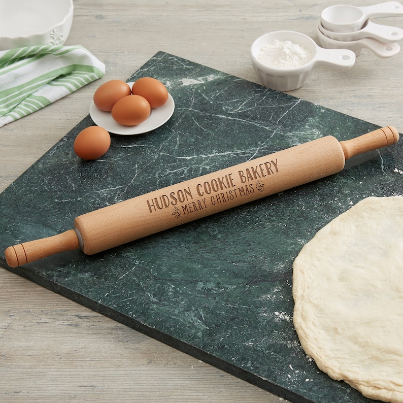 Puede incluir: Un rodillo de madera con el texto "Hudson Cookie Bakery Merry Christmas" grabado en &eacute;l. El rodillo est&aacute; sobre una superficie de m&aacute;rmol verde con una bola de masa y huevos.