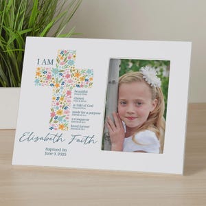 May include: White picture frame with a floral cross design and the text "I AM" and a list of religious phrases. The frame has a photo of a young girl with a white flower in her hair. The text below the photo reads "Elizabeth Faith Baptized On June 9, 2025".