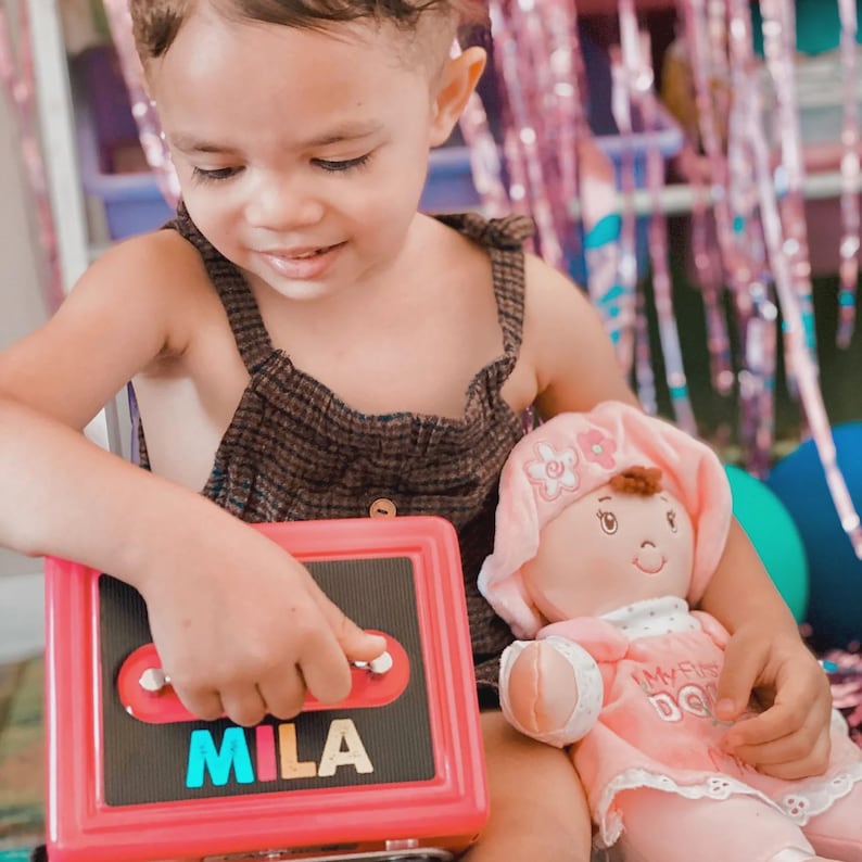 Puede incluir: Un ni&ntilde;o sostiene un juguete rosa con el nombre "MILA" en letras coloridas. El ni&ntilde;o tambi&eacute;n sostiene una mu&ntilde;eca rosa con un gorro. El ni&ntilde;o lleva un atuendo con estampado marr&oacute;n y negro.