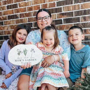 Pu&ograve; includere: Una famiglia posa con un cartello decorativo in pietra. Il cartello ovale presenta illustrazioni floreali e il testo "MOM'S GARDEN love grows here". Il cartello &egrave; tenuto da una persona e la famiglia &egrave; seduta davanti a un muro di mattoni.