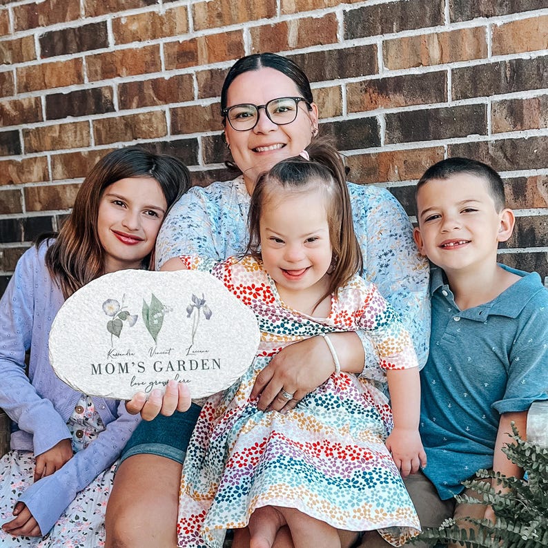Pu&ograve; includere: Una famiglia posa con un cartello decorativo in pietra. Il cartello ovale presenta illustrazioni floreali e il testo "MOM'S GARDEN love grows here". Il cartello &egrave; tenuto da una persona e la famiglia &egrave; seduta davanti a un muro di mattoni.