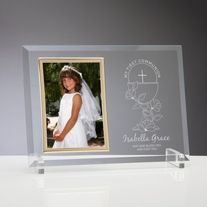 May include: A clear glass picture frame with a gold trim around the photo opening. The frame has a white etched design of a chalice with a cross and flowers. The text "My First Communion" is etched above the chalice and the text "Isabella Grace May God Bless You and Keep You" is etched below the chalice.