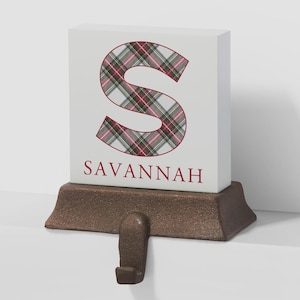 May include: A white rectangular block with a large letter S featuring a red, green, and white plaid pattern. The name SAVANNAH is printed in red below the letter. The block is mounted on a dark brown, textured cast iron stocking hook base.