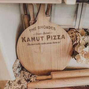 May include: A round wooden pizza peel featuring the engraved text THE RHOADES, Fresh-Baked, KAMUT PIZZA, and The best homemade pizza there is! The light-colored wood board has a long handle and is positioned near several wooden rolling pins.