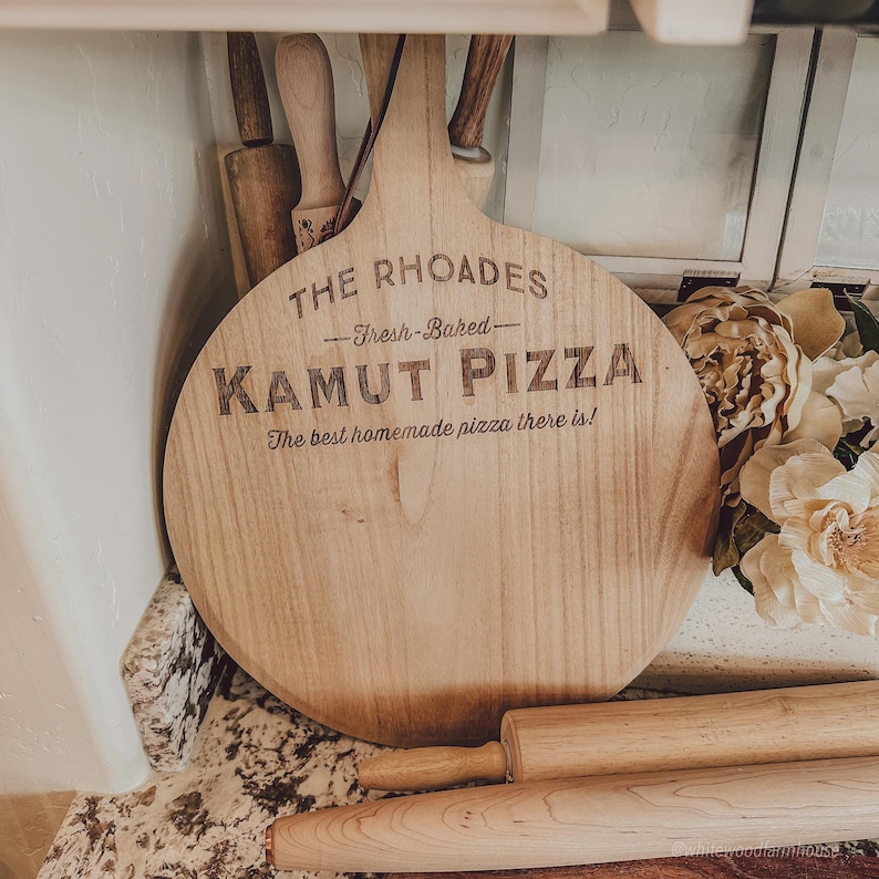 May include: A round wooden pizza peel featuring the engraved text THE RHOADES, Fresh-Baked, KAMUT PIZZA, and The best homemade pizza there is! The light-colored wood board has a long handle and is positioned near several wooden rolling pins.