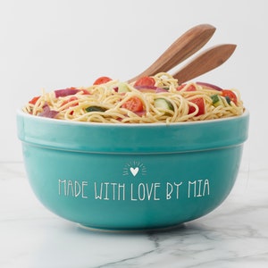 May include: A teal ceramic bowl filled with pasta salad containing cherry tomatoes, cucumber, and red onion. The bowl features the white text MADE WITH LOVE BY MIA and a small heart icon. Two wooden salad servers are tucked into the pasta.