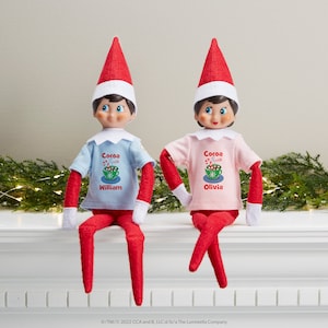 May include: Two elf dolls wearing red and white elf hats, red pants, and white gloves. The elf on the left is wearing a light blue shirt with the text "Cocoa with William" and the elf on the right is wearing a pink shirt with the text "Cocoa with Olivia".