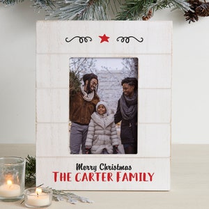 May include: White wooden picture frame with a red star and the text "Merry Christmas THE CARTER FAMILY". The frame has a photo of a family of three standing outside in the winter.
