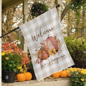 May include: A beige and white checkered house flag with the words "Welcome The Hamptons" in brown script. The flag features watercolor pumpkins and fall foliage. The flag is hanging on a black pole on a porch.