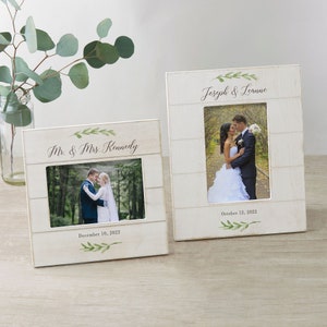 May include: Two white wooden picture frames with a rustic woodgrain finish. Each frame has a small green leafy design at the top and bottom. The frames are personalized with the names of the couples and their wedding dates. The frames are sitting on a light brown wooden surface.