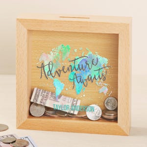 May include: A wooden shadow box with a watercolor world map design and the text "Adventure Awaits". The box has a slot on the top for inserting money. The box is filled with coins and a dollar bill.