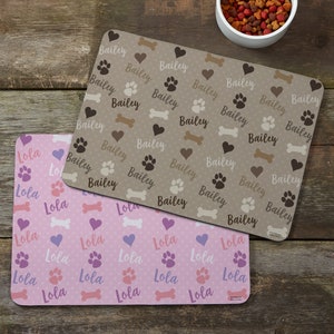 K&ouml;nnte beinhalten: Zwei Futternapfunterlagen. Eine ist beige mit Pfotenabdr&uuml;cken, Herzen und Knochenformen, und dem Namen "Bailey" wiederholt. Die andere ist rosa mit Punkten, Pfotenabdr&uuml;cken, Herzen, Knochenformen und dem Namen "Lola" wiederholt. Im Hintergrund befindet sich eine Sch&uuml;ssel mit Tierfutter.
