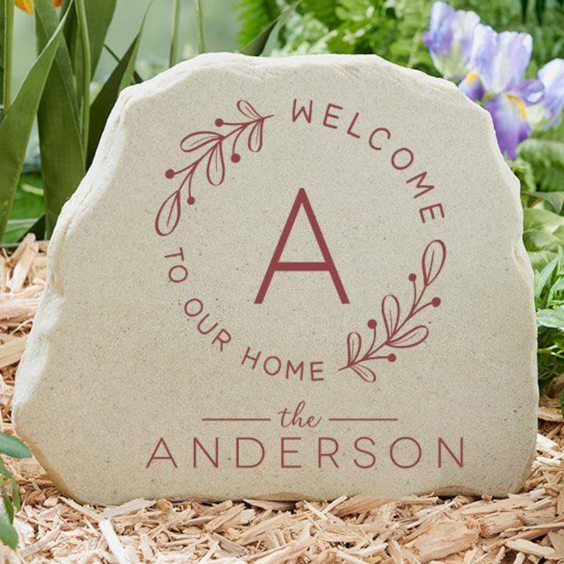 May include: A beige stone garden plaque with the words "WELCOME TO OUR HOME" in burgundy script, encircling a large letter "A". Below, the family name "the ANDERSON" is printed. The plaque is surrounded by greenery.