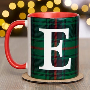Puede incluir: Una taza roja y blanca con un patr&oacute;n de cuadros verdes y la letra "E" en blanco. La taza est&aacute; sobre un posavasos marr&oacute;n.