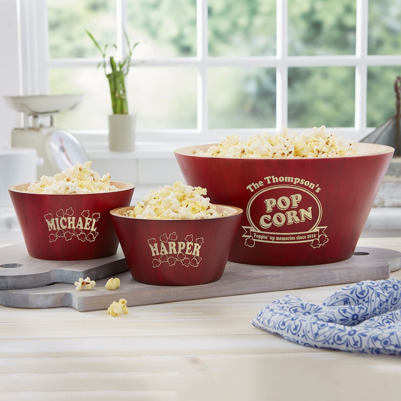 May include: Three red wooden popcorn bowls with personalized names. The largest bowl says "The Thompson's Pop Corn Poppin' up memories since 2018". The medium bowl says "Harper" and the smallest bowl says "Michael".