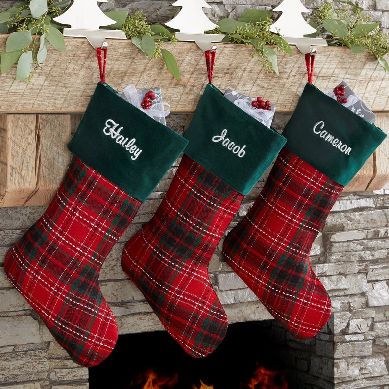 May include: Three Christmas stockings with red and green plaid patterns. Each stocking has a dark green cuff with a name embroidered in white: Hailey, Jacob, and Cameron. The stockings are hanging from a wooden mantel.
