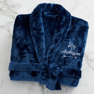 May include: A plush navy blue robe with a shawl collar and a tie belt. The robe features the embroidered text "Mr. Anderson" with an arrow. The soft, textured fabric suggests comfort and warmth. The robe is folded neatly on a marble surface.