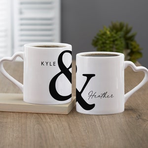 May include: Two white ceramic mugs with heart-shaped handles, filled with coffee. One mug has the name "Kyle" and an ampersand, the other has "Heather" and an ampersand. The mugs are on a wooden surface.
