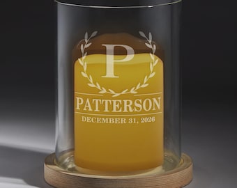 May include: A cylindrical glass hurricane candle holder with a light wood base containing a yellow pillar candle. The candle features an etched design with a laurel wreath, the letter P, the name PATTERSON, and the date DECEMBER 31, 2026.