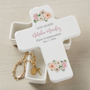 May include: White ceramic cross-shaped box with a floral design and the text "God Bless Natalia Mendez First Communion May 7, 2024". The box is open and a gold rosary is inside.