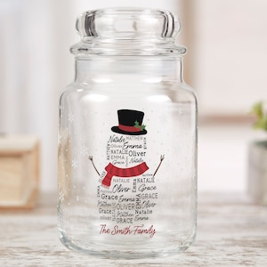 May include: A clear glass jar with a snowman design. The snowman is made of white snowflakes and has a black top hat, red scarf, and green holly. The jar is decorated with the names "Natalie, Matthew, Oliver, Emma, Grace" and "The Smith Family".