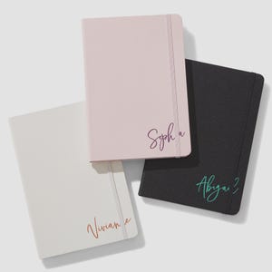 May include: Three rectangular notebooks with elastic closure bands. One is light pink with the name Sophia in dark purple script, one is white with the name Vivian in orange script, and one is black with the name Abigail in teal script.