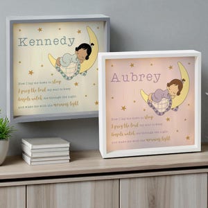 May include: Two framed nursery art pieces. One is gray, the other white. Each features a sleeping baby on a crescent moon, surrounded by stars. The text reads "Kennedy" and "Aubrey" with a prayer.