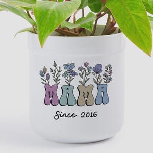 May include: A white ceramic plant pot containing a green leafy plant. The pot features a graphic design with the word MAMA in pastel-colored, wavy-shaped letters, each topped with illustrated wildflowers. The text Since 2016 is printed below.