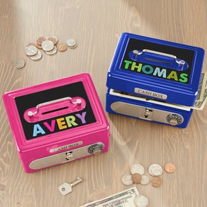 Puede incluir: Dos cajas de efectivo personalizadas, una rosa y otra azul, sobre una superficie de madera. La caja rosa tiene el nombre "AVERY" en letras coloridas, mientras que la caja azul dice "THOMAS". Ambas cajas tienen una ranura para el dinero, una cerradura de combinaci&oacute;n y la inscripci&oacute;n "CASH BOX".
